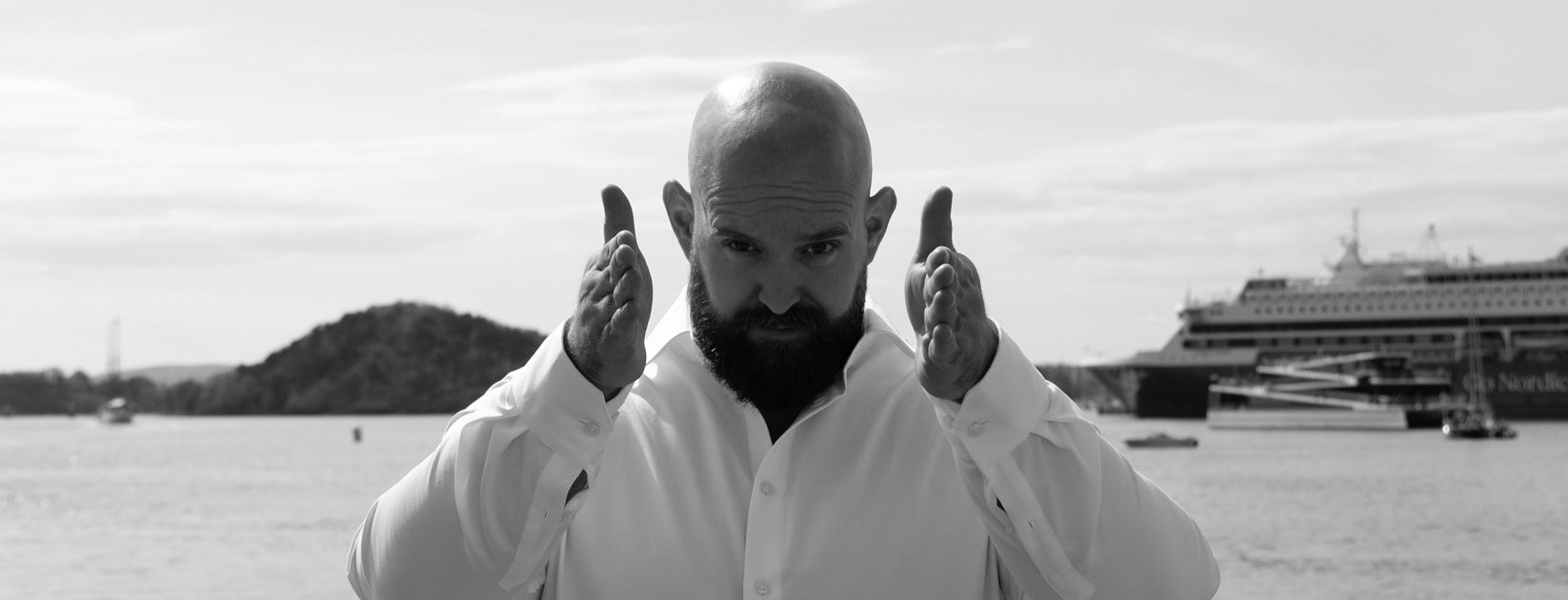 Peter R. Valderhaug i hvit skjorte ved sjøen med hendene løftet foran seg.